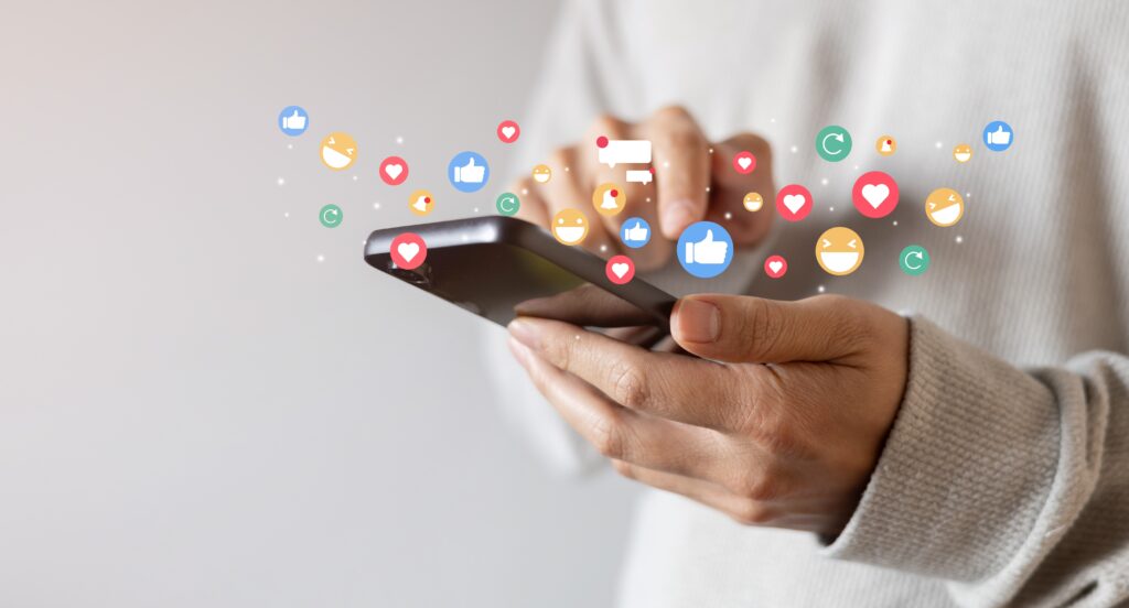 Primer plano de las manos de una persona usando un teléfono celular del que brotan íconos flotantes de reacciones como likes, corazones y emojis, representando la dopamina digital y la necesidad de validación en redes sociales.